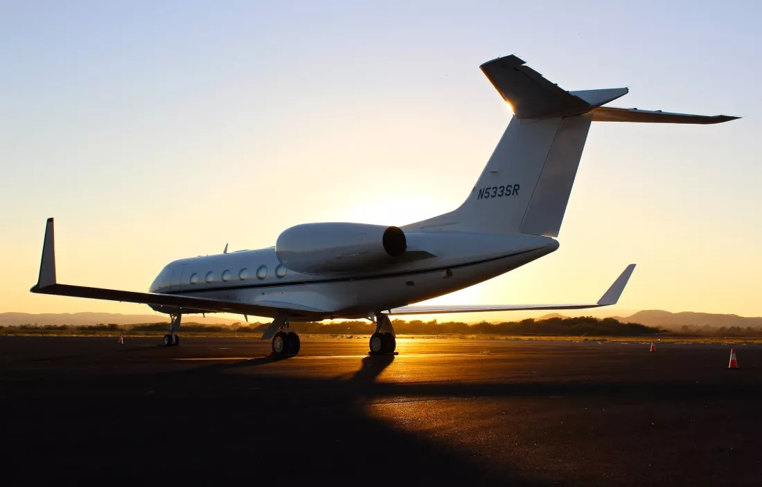 Avión privado estacionado en la pista al atardecer, con el sol poniéndose detrás del fuselaje. El cielo está despejado y el paisaje muestra colinas en el fondo, con conos naranjas visibles en el suelo cerca de la aeronave.
