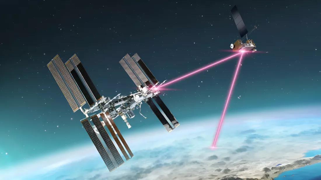 La imagen muestra una ilustración de la Estación Espacial Internacional (ISS) en el espacio, comunicándose con un satélite mediante un enlace láser visible como haces de luz rosada. La Tierra se ve en el fondo, mostrando una curvatura sutil que resalta la posición orbital de ambos objetos.