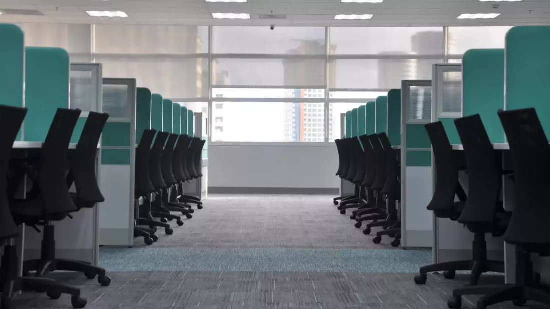 Oficina vacía con una fila de escritorios con separadores de color turquesa y sillas negras a ambos lados, iluminada por luz natural que entra a través de grandes ventanales al fondo.