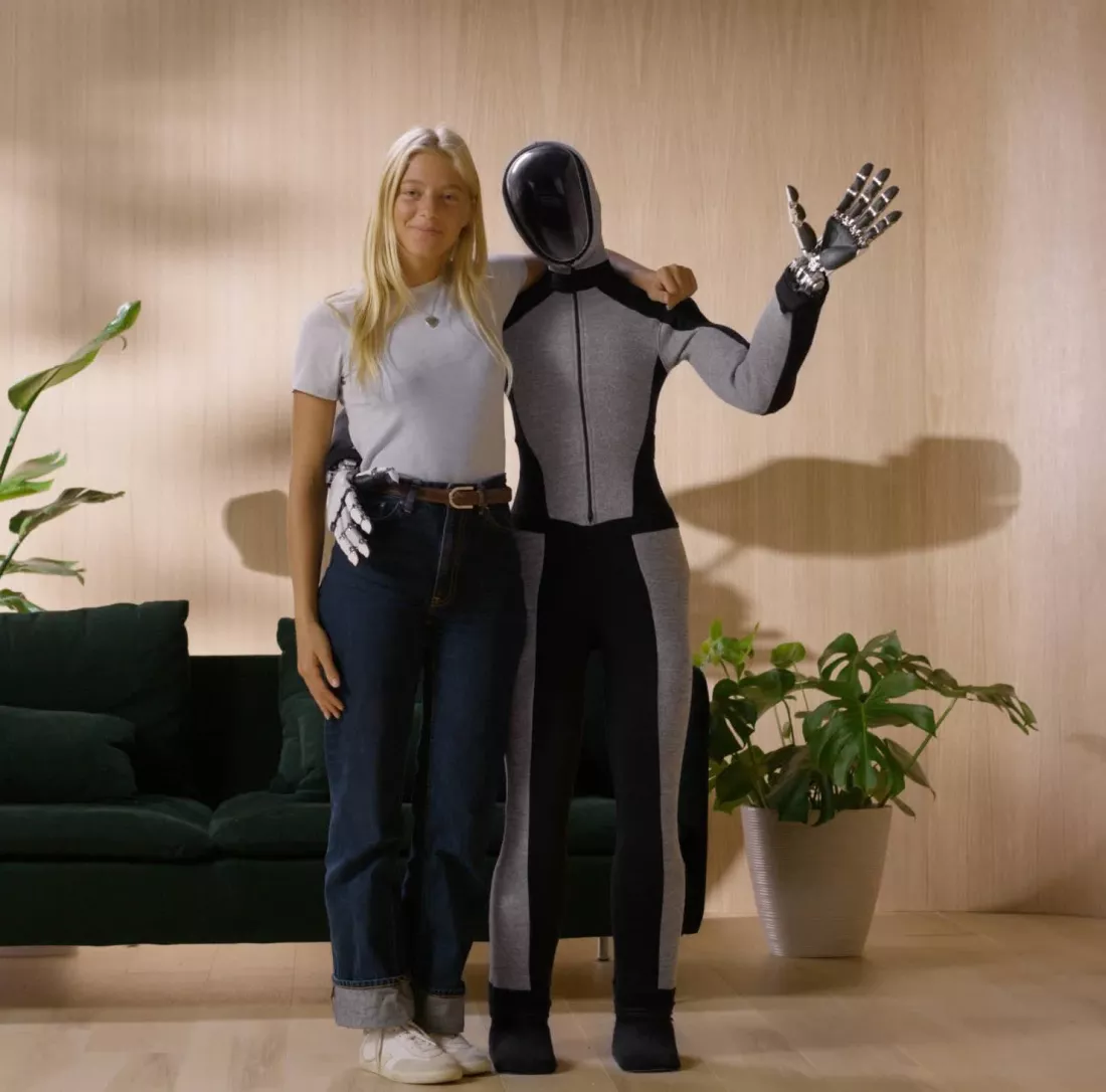 Una joven rubia posando junto a un robot humanoide de tamaño similar. El robot, de aspecto futurista, tiene un diseño sin rostro y está vestido con un traje ajustado de dos tonos, gris y negro. La joven tiene su brazo alrededor del robot, que levanta una mano en un gesto de saludo. La escena se desarrolla en un entorno hogareño con paredes de madera clara, un sofá verde oscuro y una planta decorativa en el fondo, lo que sugiere un ambiente cómodo y moderno.